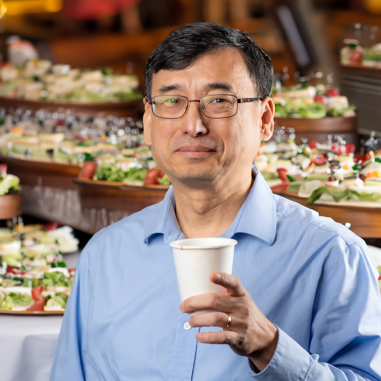 Chengping Chang holding up a cup of coffee with sandwiches in the background