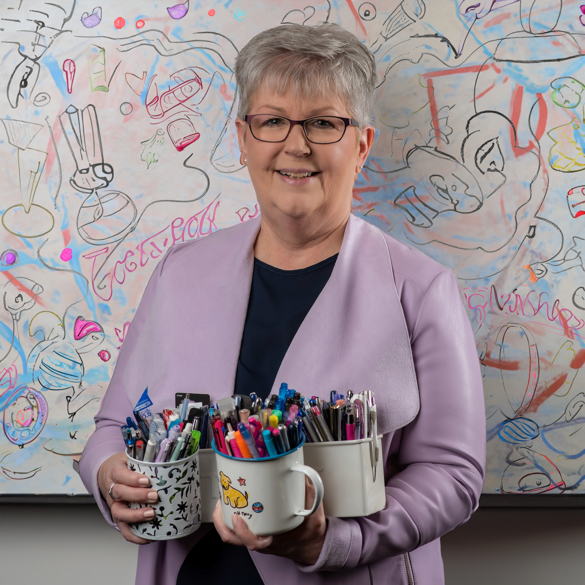 Mary Ann friedman fun photo posing with mugs full of markers and pens with a dry erase board full of doodles in the background