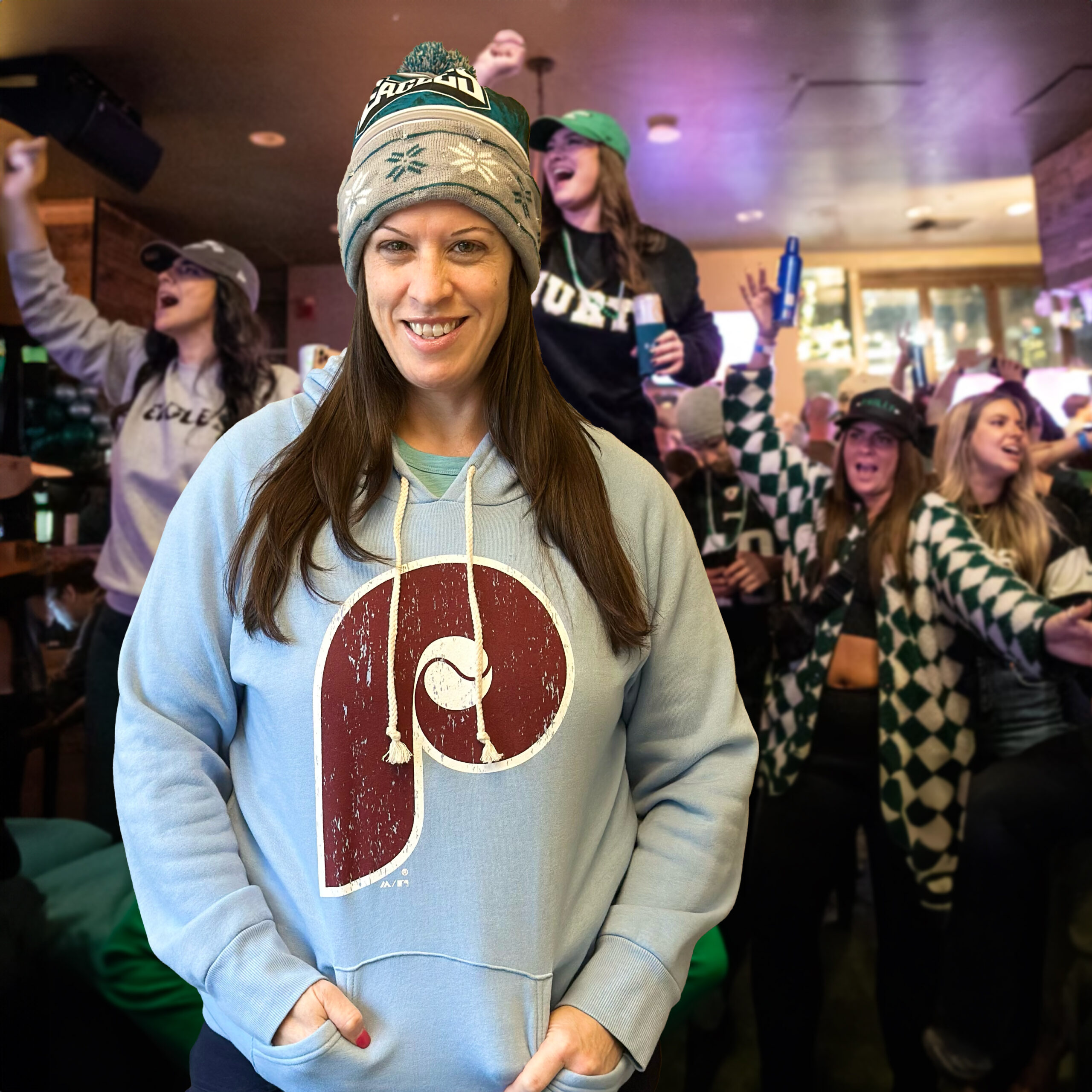 Kim Gatti posing in a Phillies hoodie and Eagles hat with Eagles fans celebrating in the background