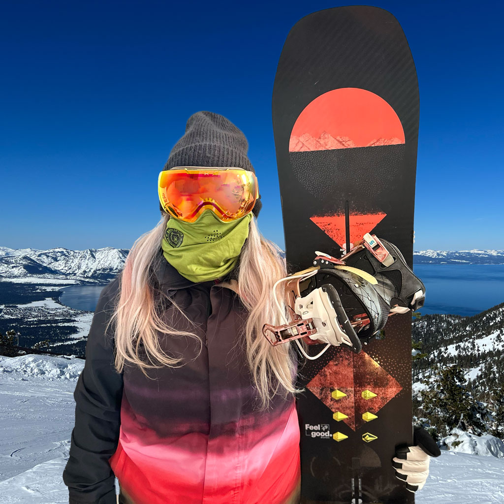 Stacy Moorhead posing with a snowboard and goggles with snow covered Lake Tahoe in the backgroud