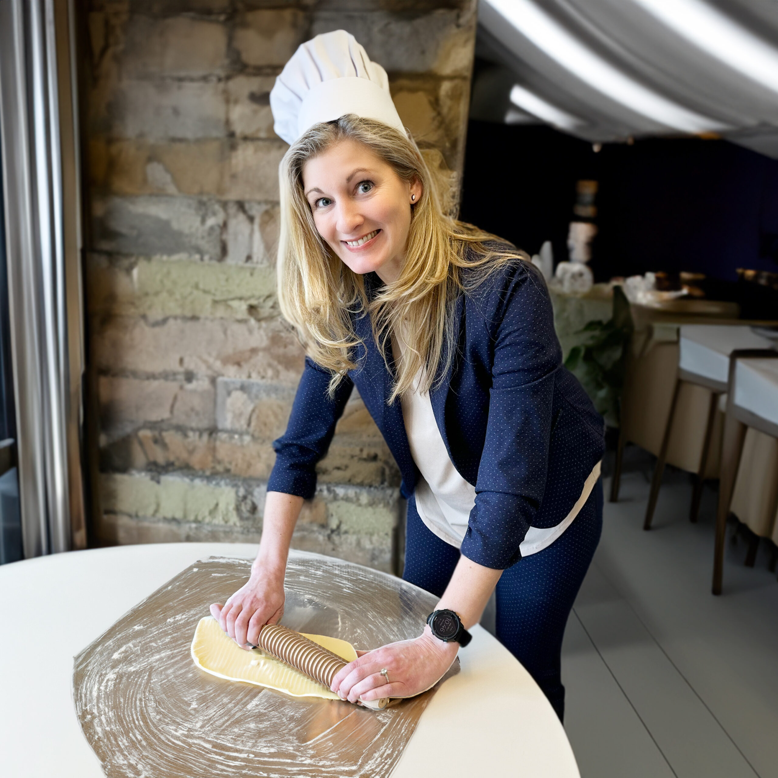 Caitlin Pletcher wearing a chef's hat rolling out dough on a table with a kitchen in the background