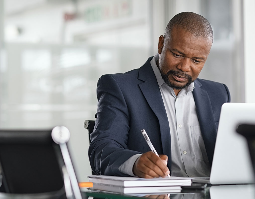 Mature business man writing his strategy on notebook while using laptop in modern office