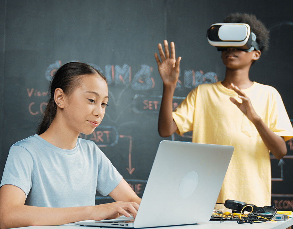 Two students in, one with a laptop, the other with a VR mask on 