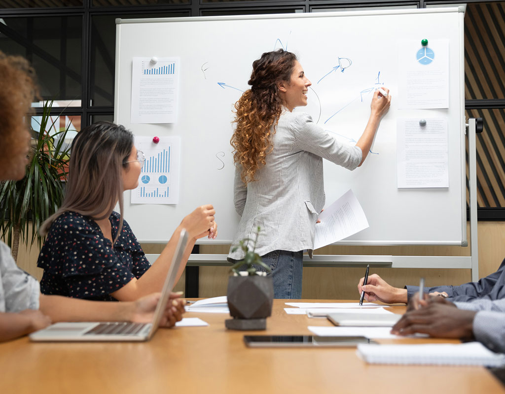 group of adults sitting at a table looking at a person drawing on a white board