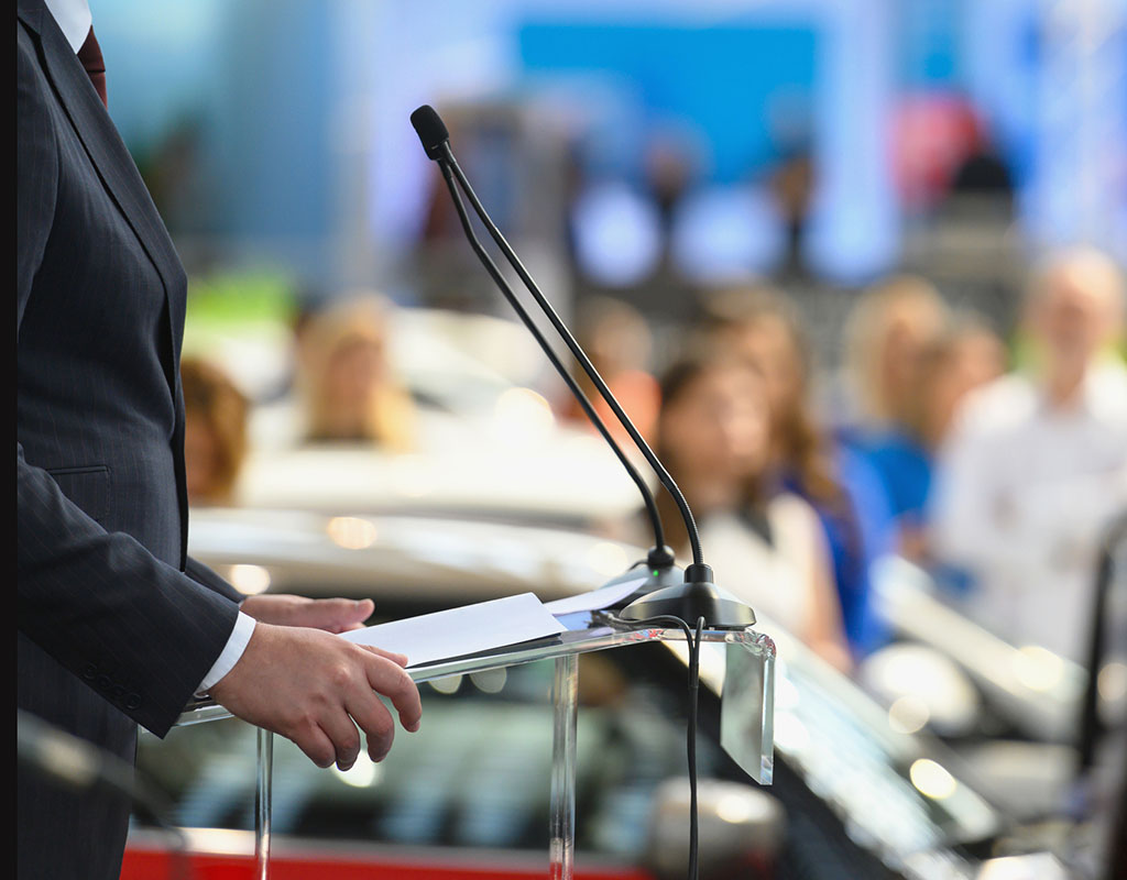 person standing at a podium with a microphone with a crowed blurred in the background