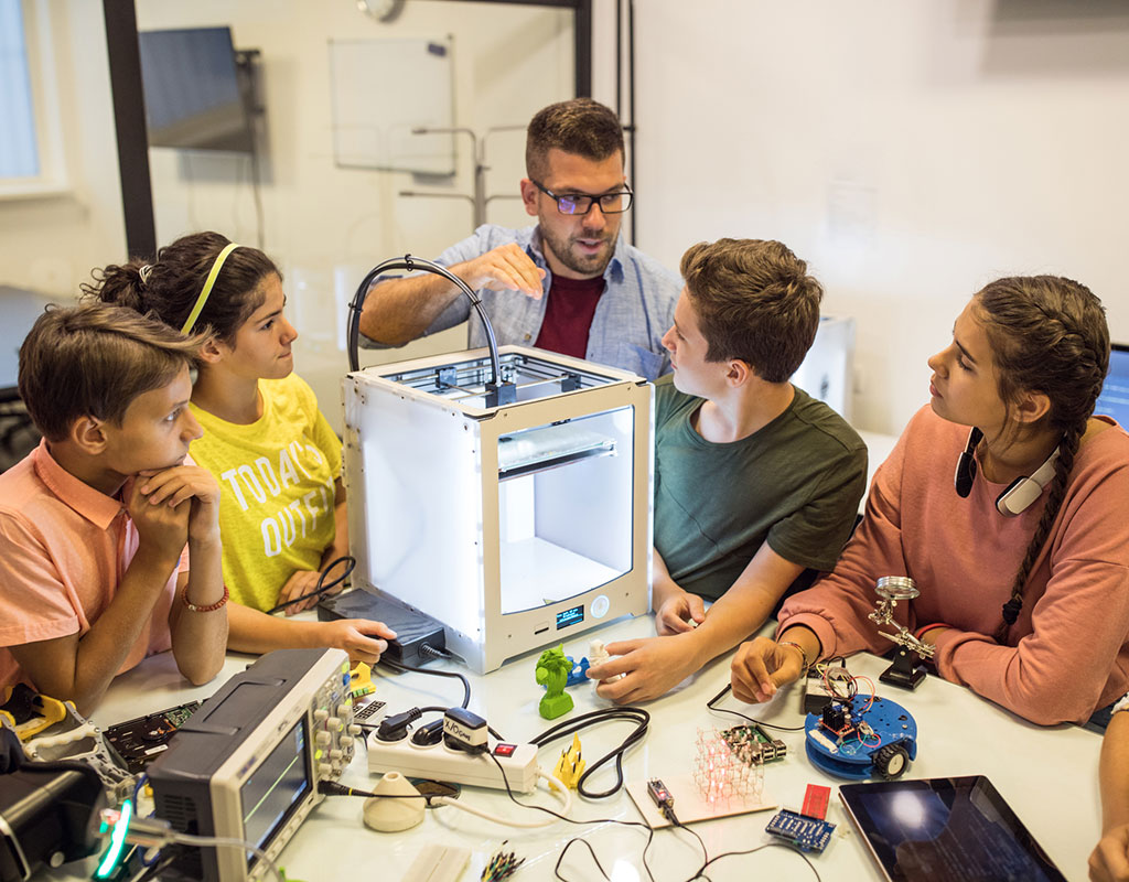 Students and teacher in a classroom gathered around a 3D printer