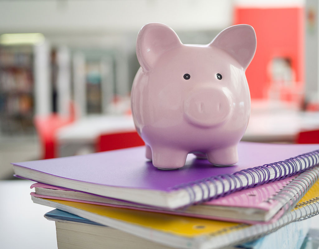 Closeup of notebooks on a desk in a classroom with a piggy bank on top