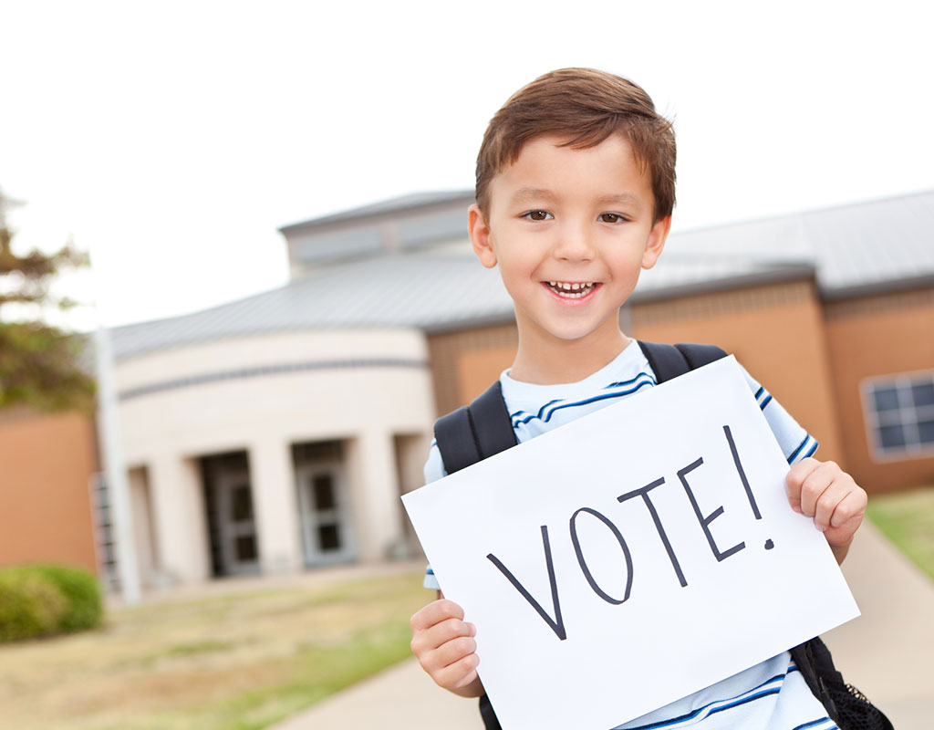 Male student standing outside a school with a backpack holding a sign that says "vote"