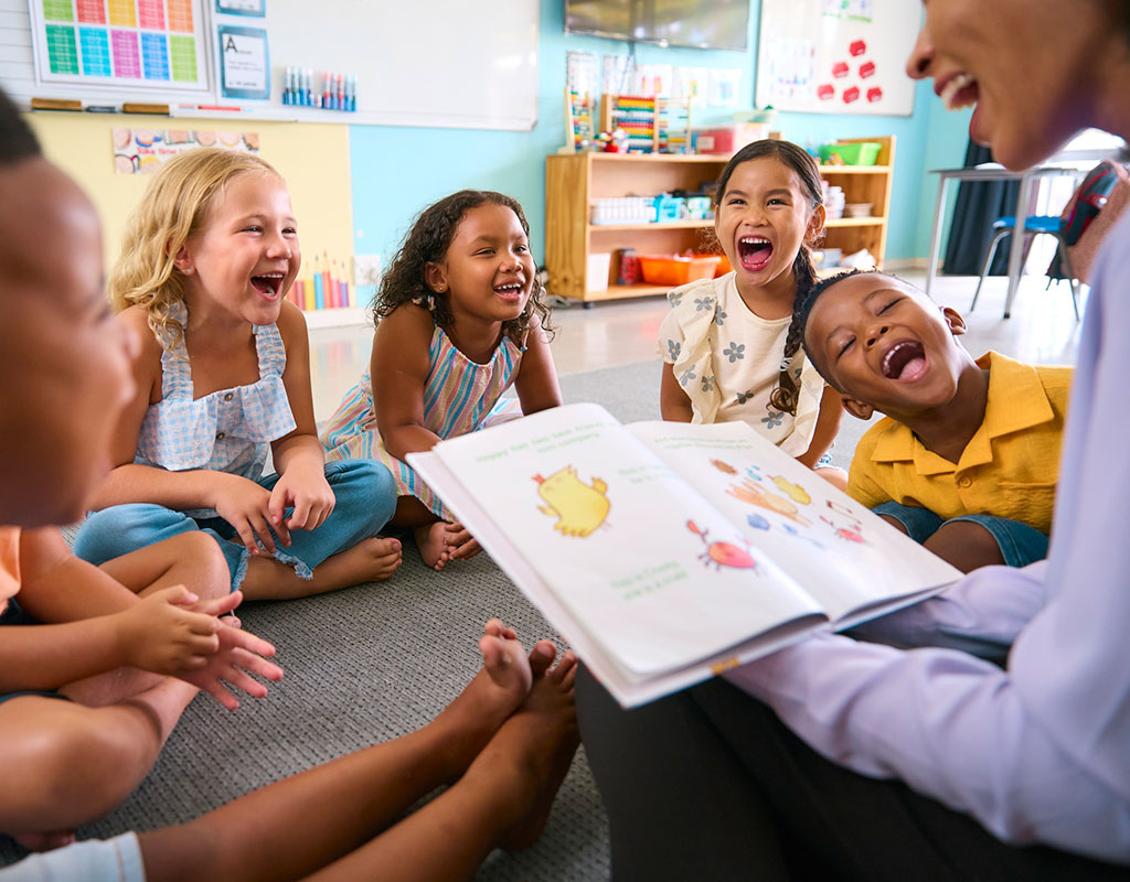 Photo of smiling children in a classroom sitting in a circle around a teach reading