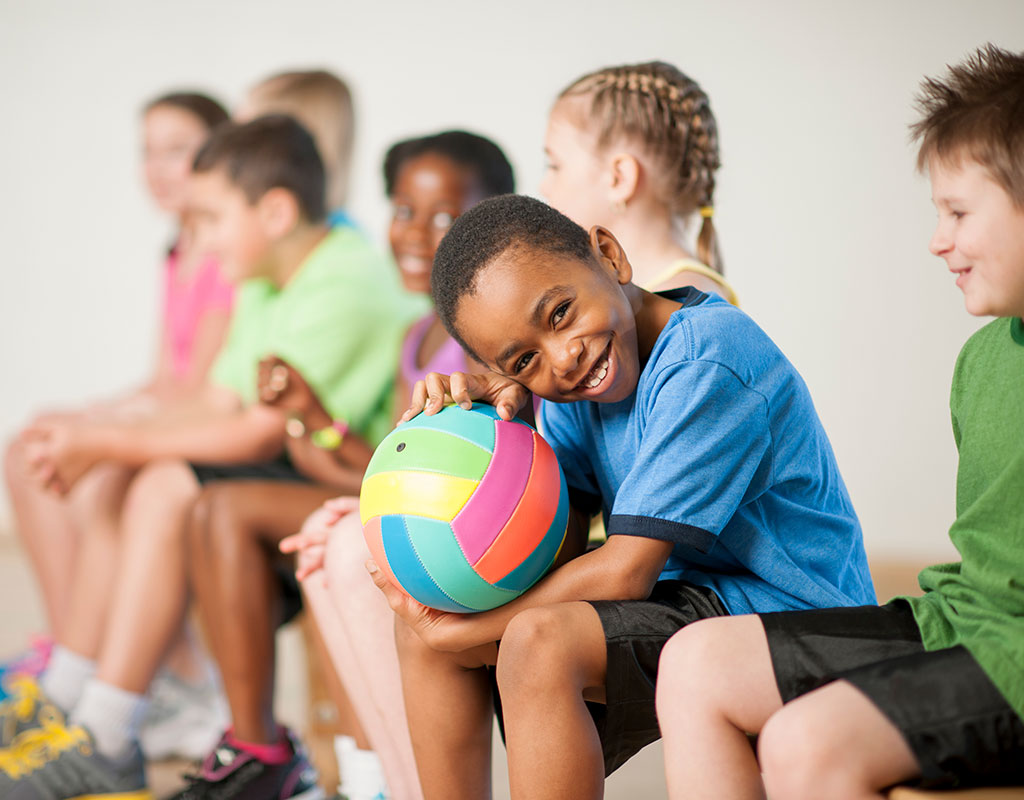 Photo of smiling children sitting on the sidelines of a volley ball gym class holding a colorful volleyball 